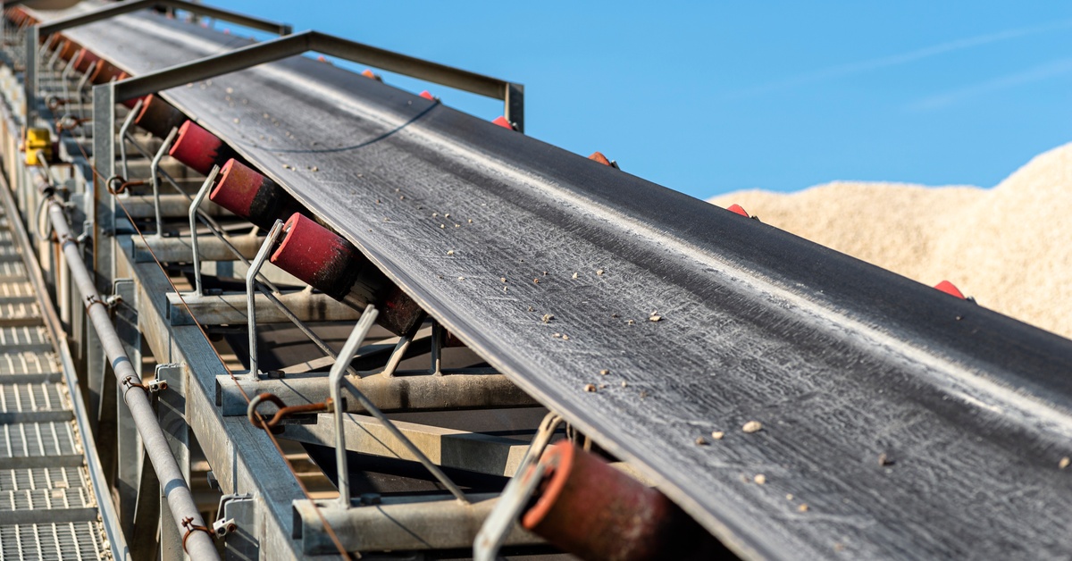 A rubber belt stretches up through a conveyor system. It features red transport rollers and a metal stairway.