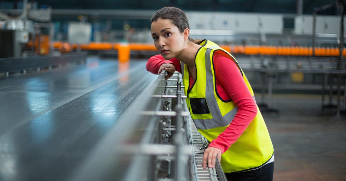 A worker in a factory examines the belt on a conveyor system. She wears a yellow safety vest as she works.