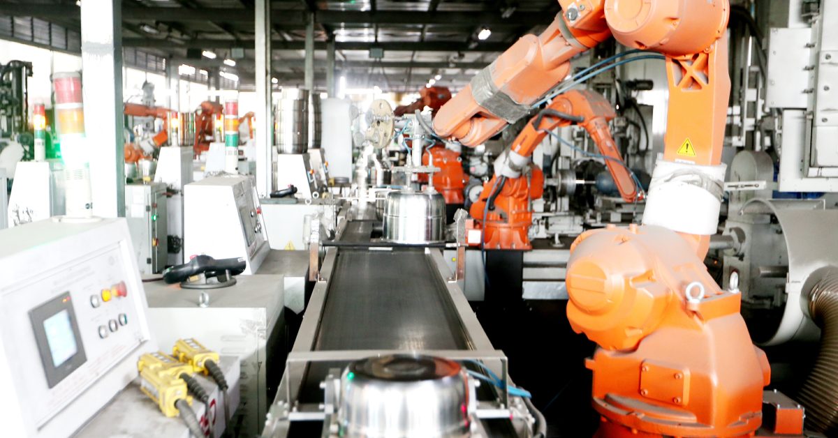 Large orange industrial machines operate in an assembly line in a factory. They sit next to a conveyor system.