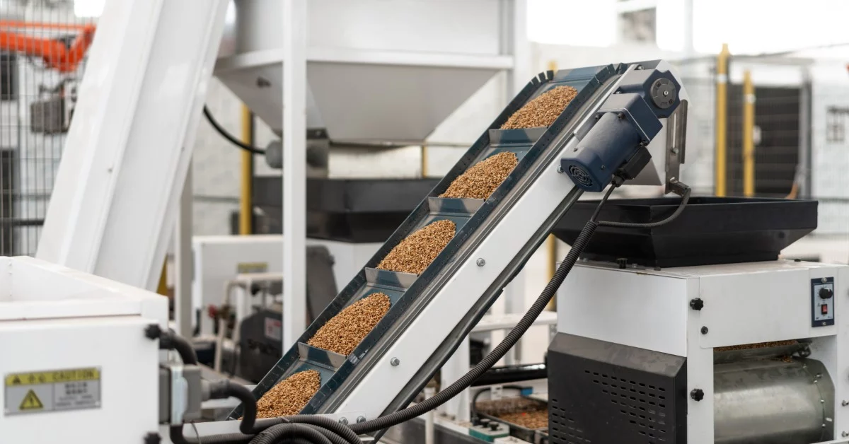 An incline conveyor moves loads of granular material inside a manufacturing facility. The interior features white walls.