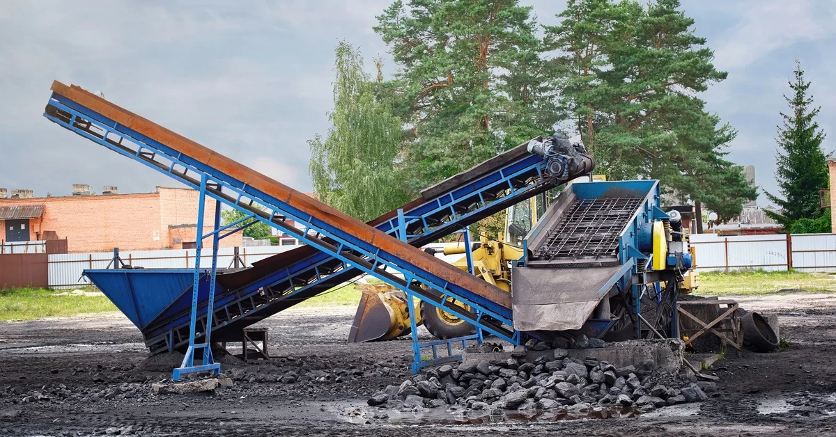Two portable belt conveyors sit next to a piece of machinery in an outdoor mining operation. Trees and a building sit nearby.